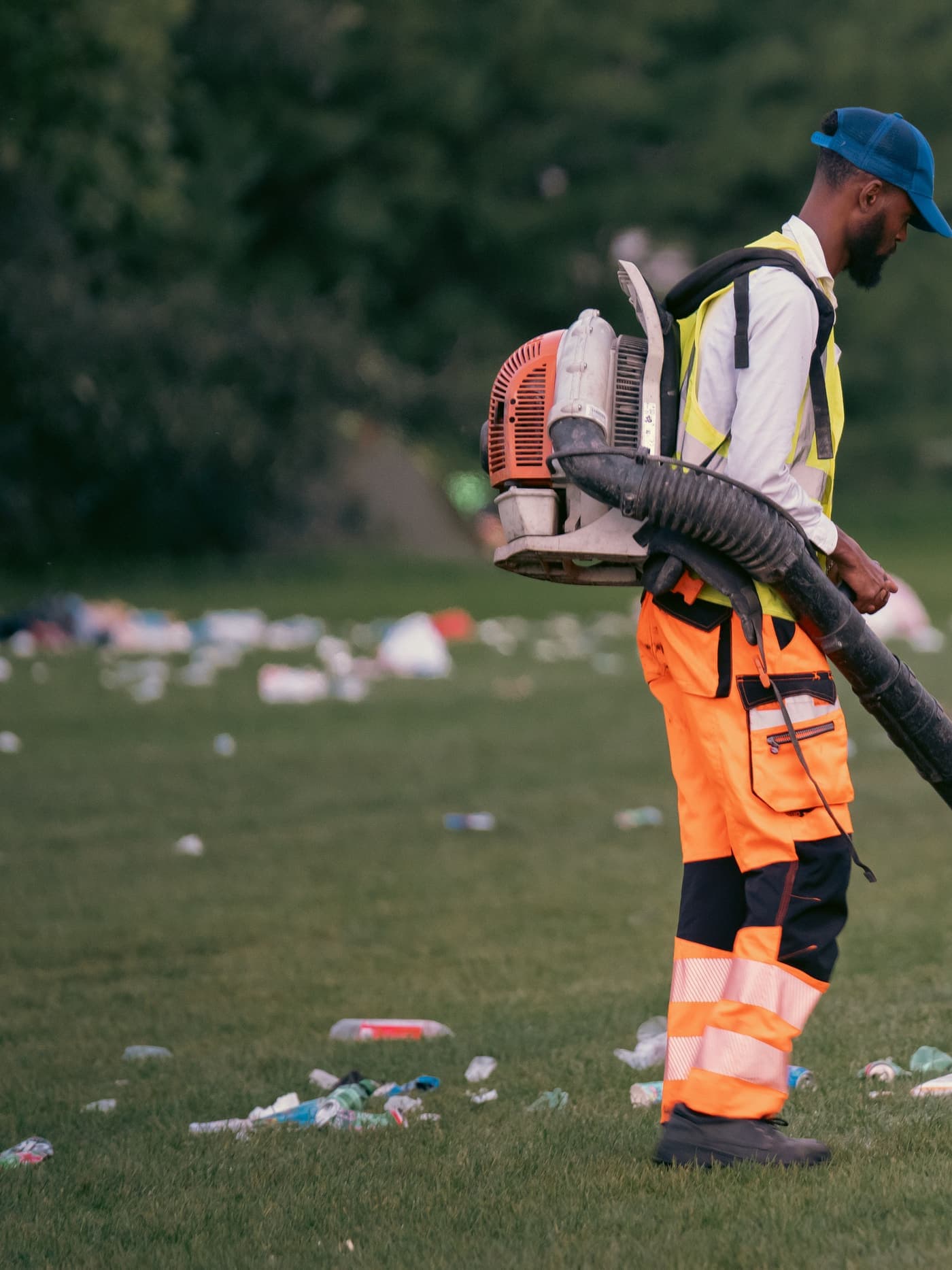 A worker in high-visibility gear clearing debris from a grassy commercial-area lawn with a backpack blower